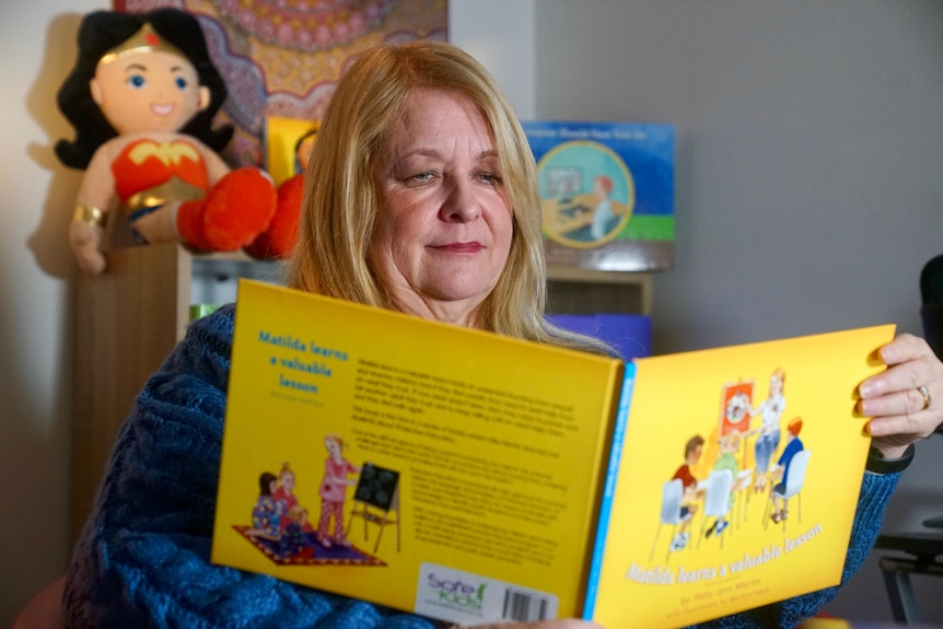 A woman sits in front of a bookshelf reading a childrens book