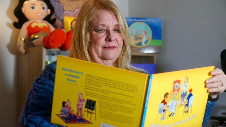 A woman sits in front of a bookshelf reading a childrens book