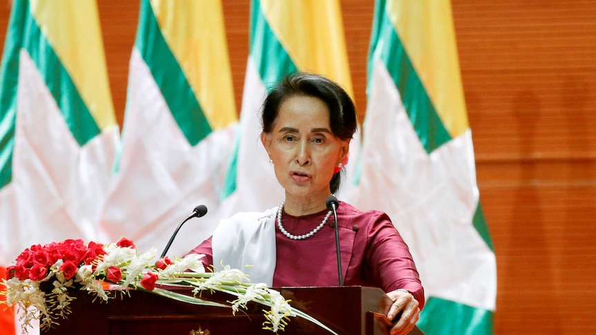 Aung San Suu Kyi speaks at a podium with flags in the background.