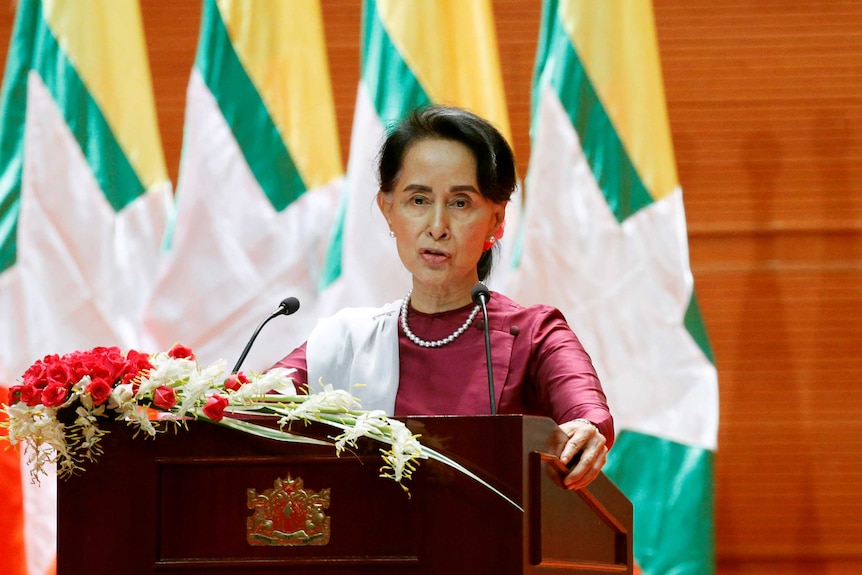 Aung San Suu Kyi speaks at a podium with flags in the background.