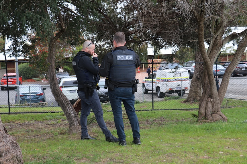 Two police officers stand chatting near a fence, with a police vehicle visible in the background.