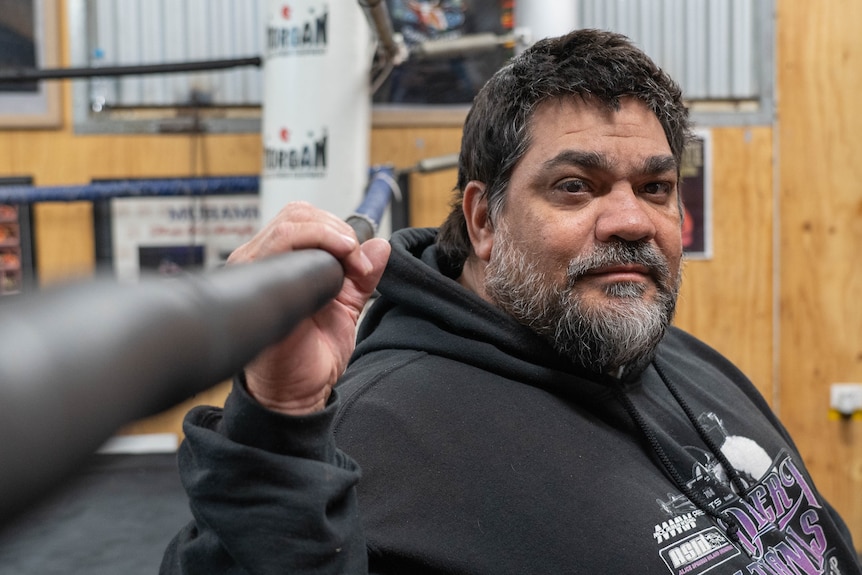 An man with dark hair and a salt-and-pepper beard, wearing a hoodie, sits in a gym.