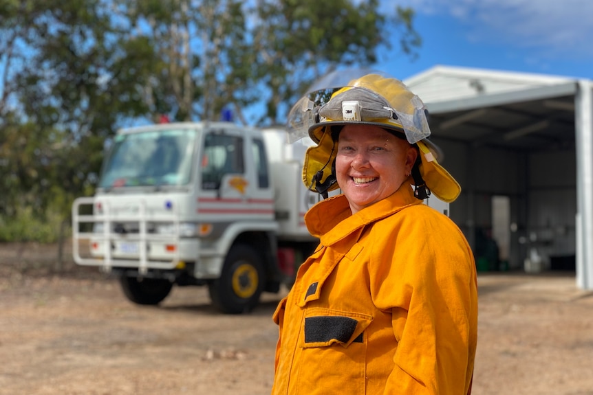 Woman in orange overalls and white helmet in foreground smiling at camera,front of fire truck and shed in bush setting