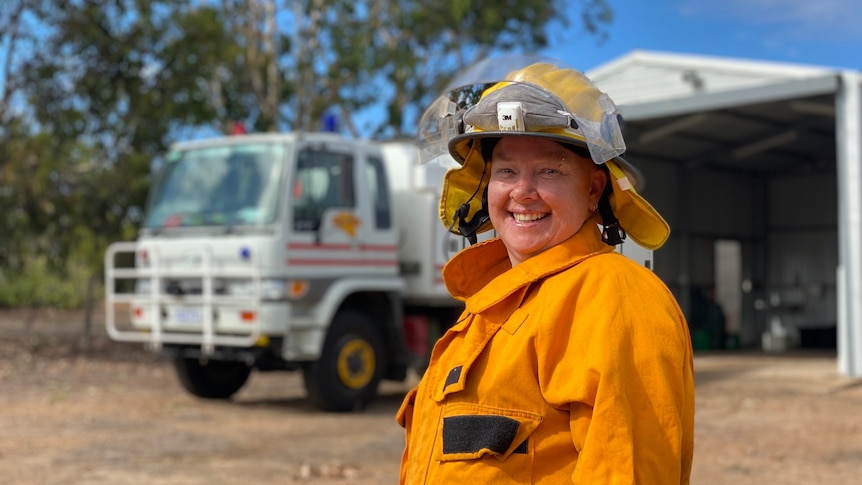 Woman in orange overalls and white helmet in foreground smiling at camera,front of fire truck and shed in bush setting