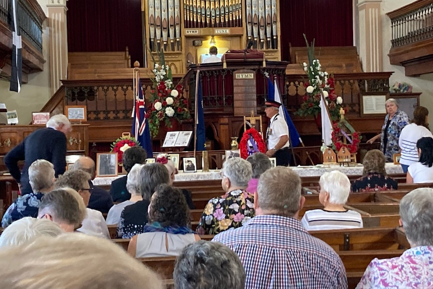 Organ pipes dominate  a building with wooden pews, three Australian flags in front, a man in uniform and people sitting.