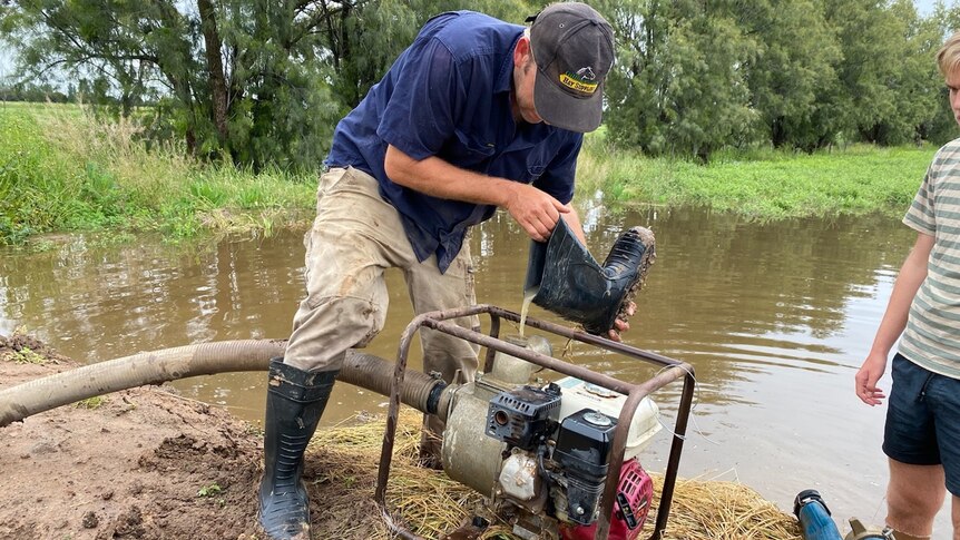 A man pours water from a gumboot into a pump. He is in a flooded area.
