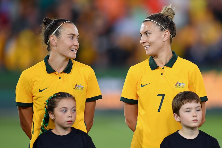 Two soccer players wearing yellow and green jerseys look at each other with two children standing in front of them