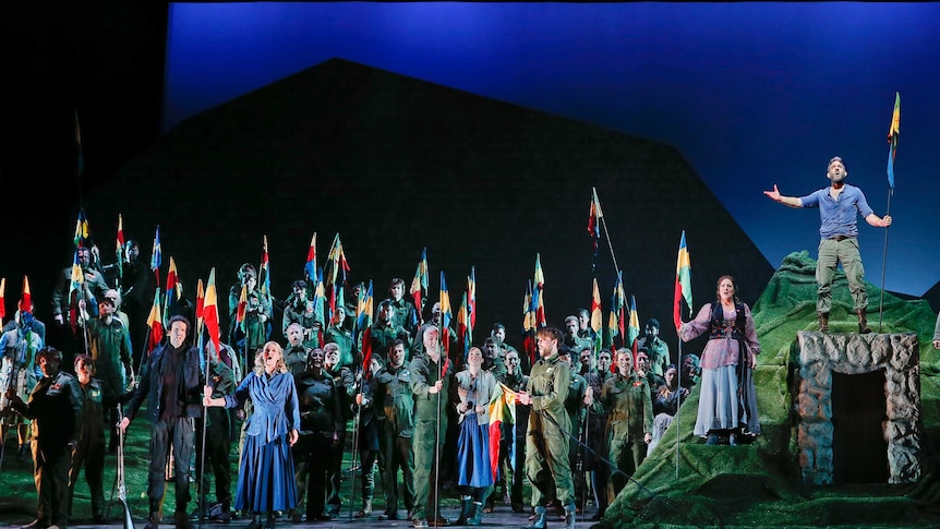 A large number of opera performers holding guns or flags stand on a stage set depicting rolling hills.
