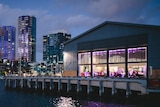 A group of revellers can be seen through the large windows of a shed on Central Pier, illuminated by purple lighting at night.