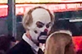 A man in a suit and clown mask stares at a blonde woman