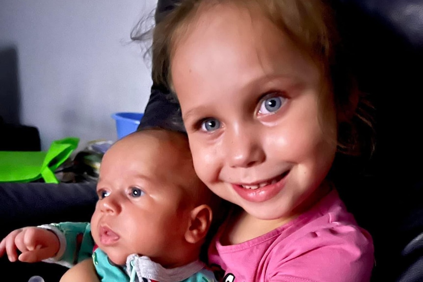 Little girl smiling while holding an infant and sitting down.