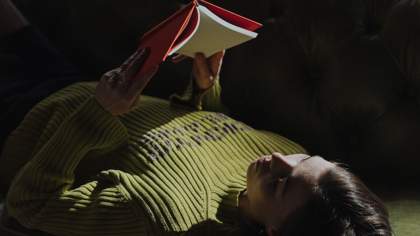 A dark, moody photo of a woman wearing a green sweater laying on her back, reading a book