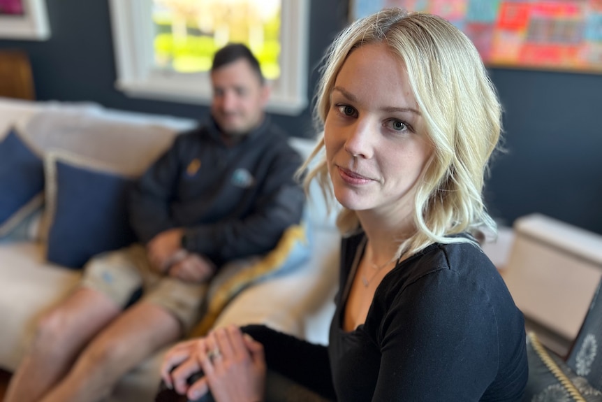 A young blonde woman looks at us on the right, a blurry man sits behind her on the couch