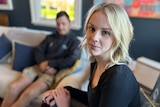 A young blonde woman looks at us on the right, a blurry man sits behind her on the couch
