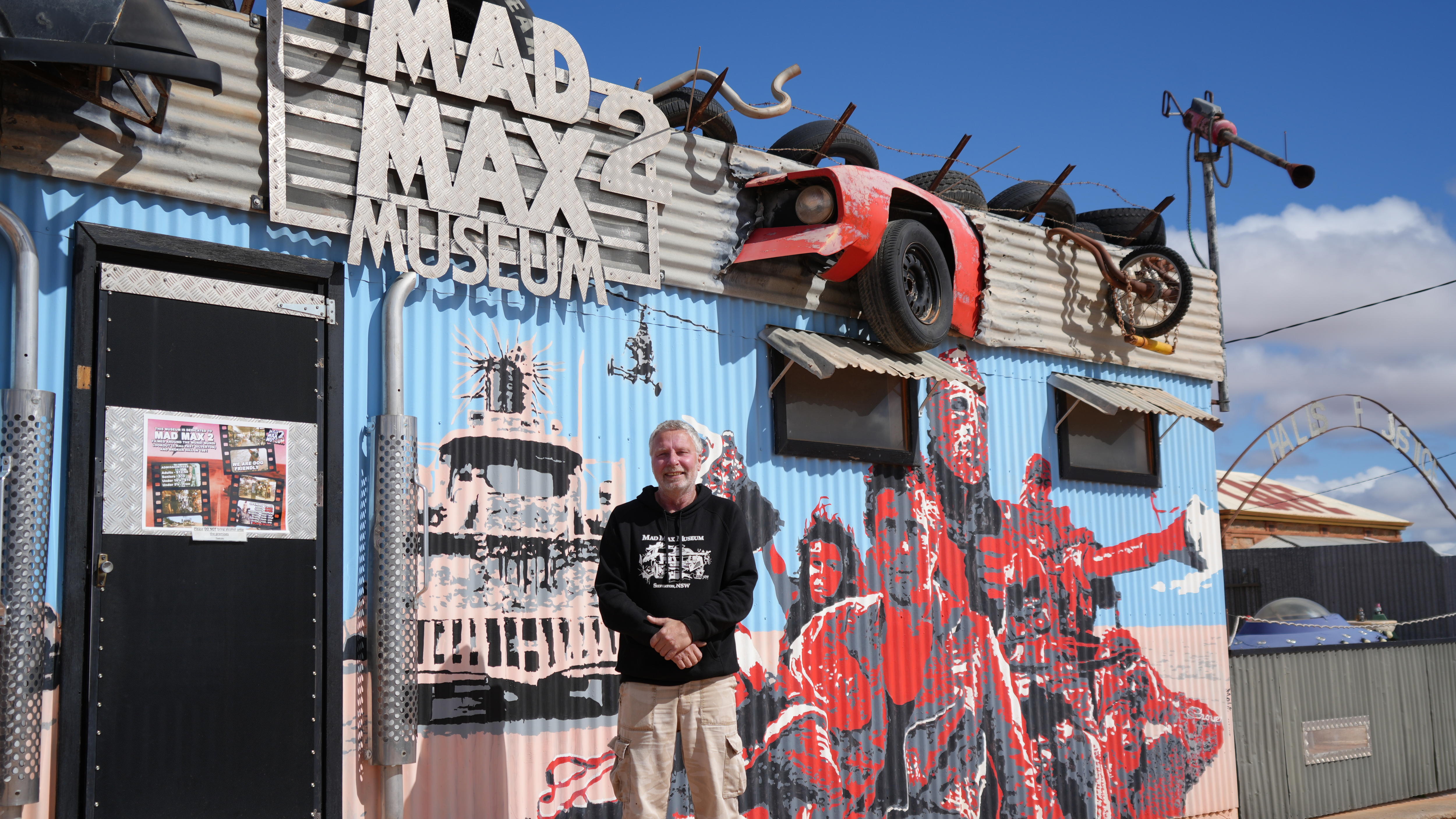 A middle-aged man wearing a black hoodie and pants standing in front of a painted metal shed with a sign saying mad max 2 museum