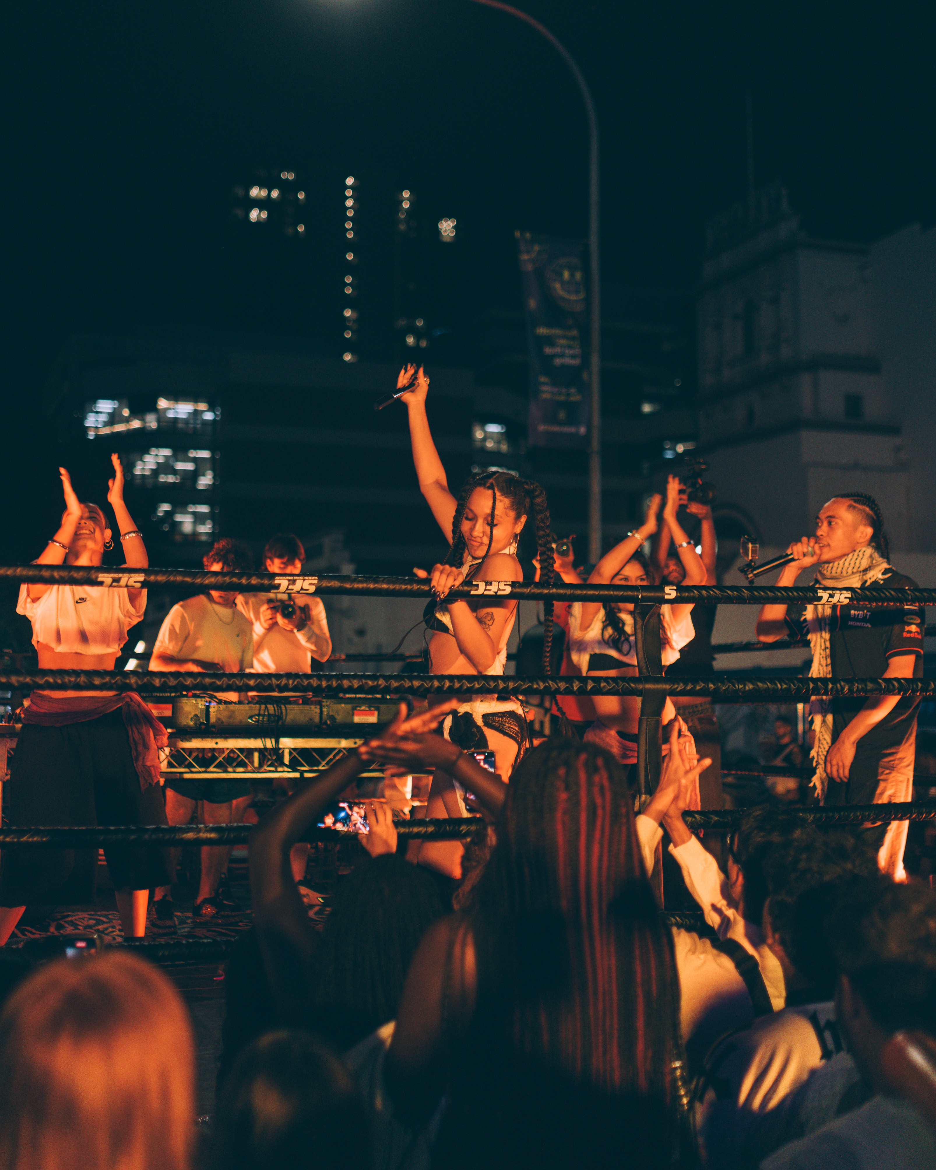 A group of young people dance and sing in an elevated boxing ring at night in front of a crowd
