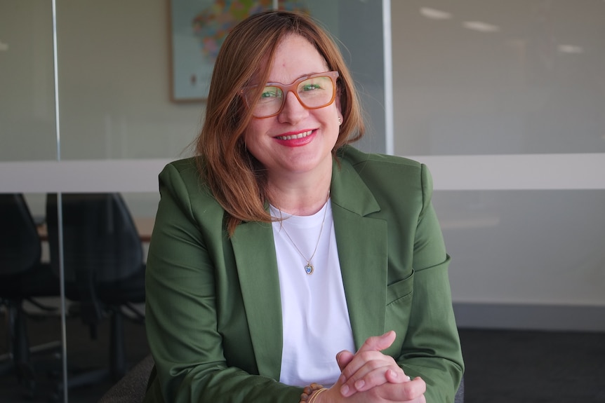 Nicole wears green blazer and orange-rimmed glasses and smiles with her hands clasped together sitting in a boardroom. 