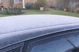 A black car parked in a front yard is frosted over on its roof and windows