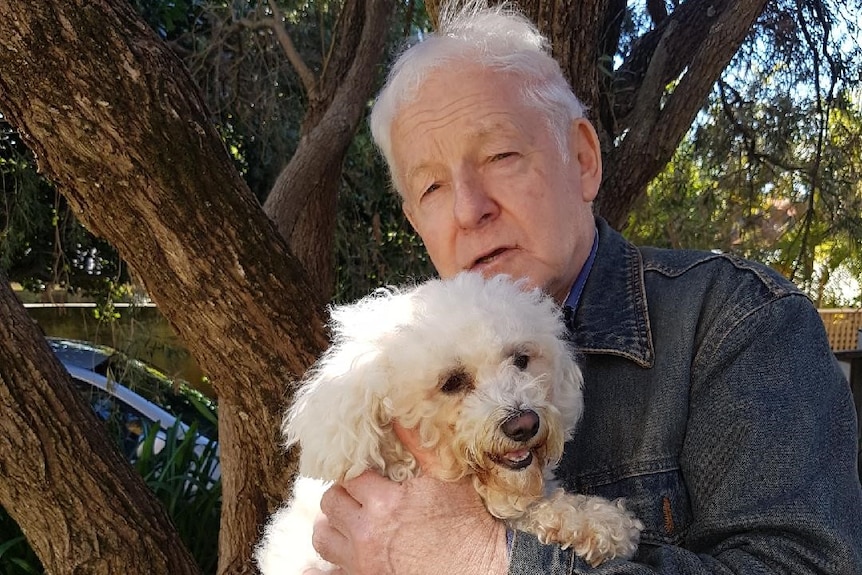 Former prisoner John Killick stands in front of a tree in a garden holding a dog.