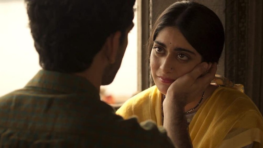 A woman with dark hair wearing a yellow sari stares at a man in profile against a window.