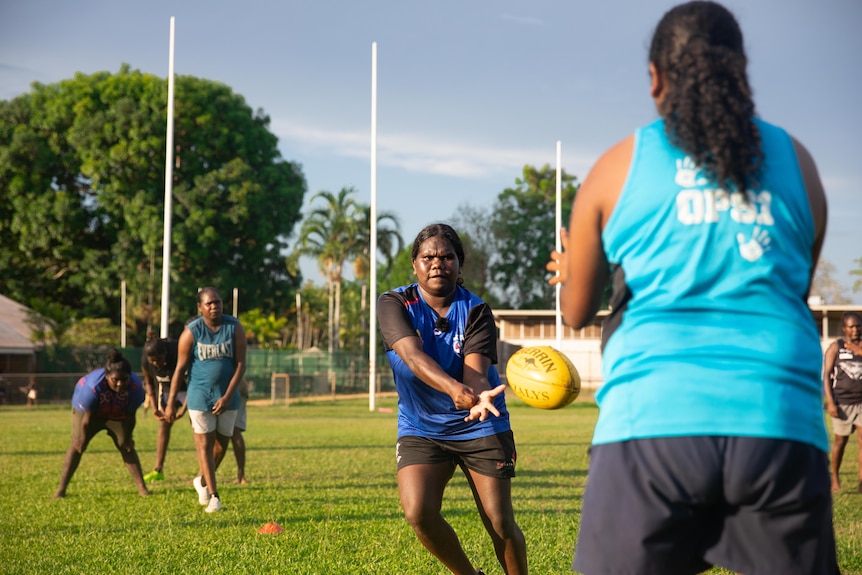 A woman in a blue shirt handballs a yellow football on an Australian Rules oval