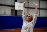 An older woman wearing a grey jumper and sunglasses has her arms outstretched above her head.