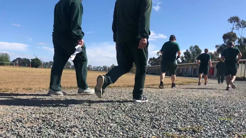 Faceless prisoners in green athletic gear walk and run along gravel track inside prison