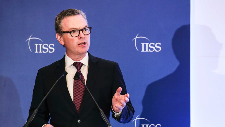 Defence minister Christopher Pyne gesticulates stage-right in front of a blue and white backdrop that reads 'IISS'.