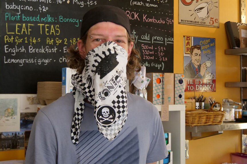 A barista wearing a black chef's cap and white and black face mask stands in a coffee shop.