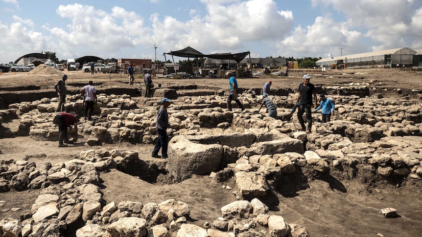 A group of archaeologists stand on and dig out large piles of rock and soil.