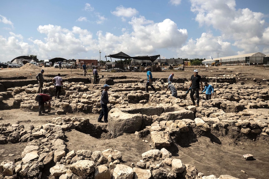 A group of archaeologists stand on and dig out large piles of rock and soil.