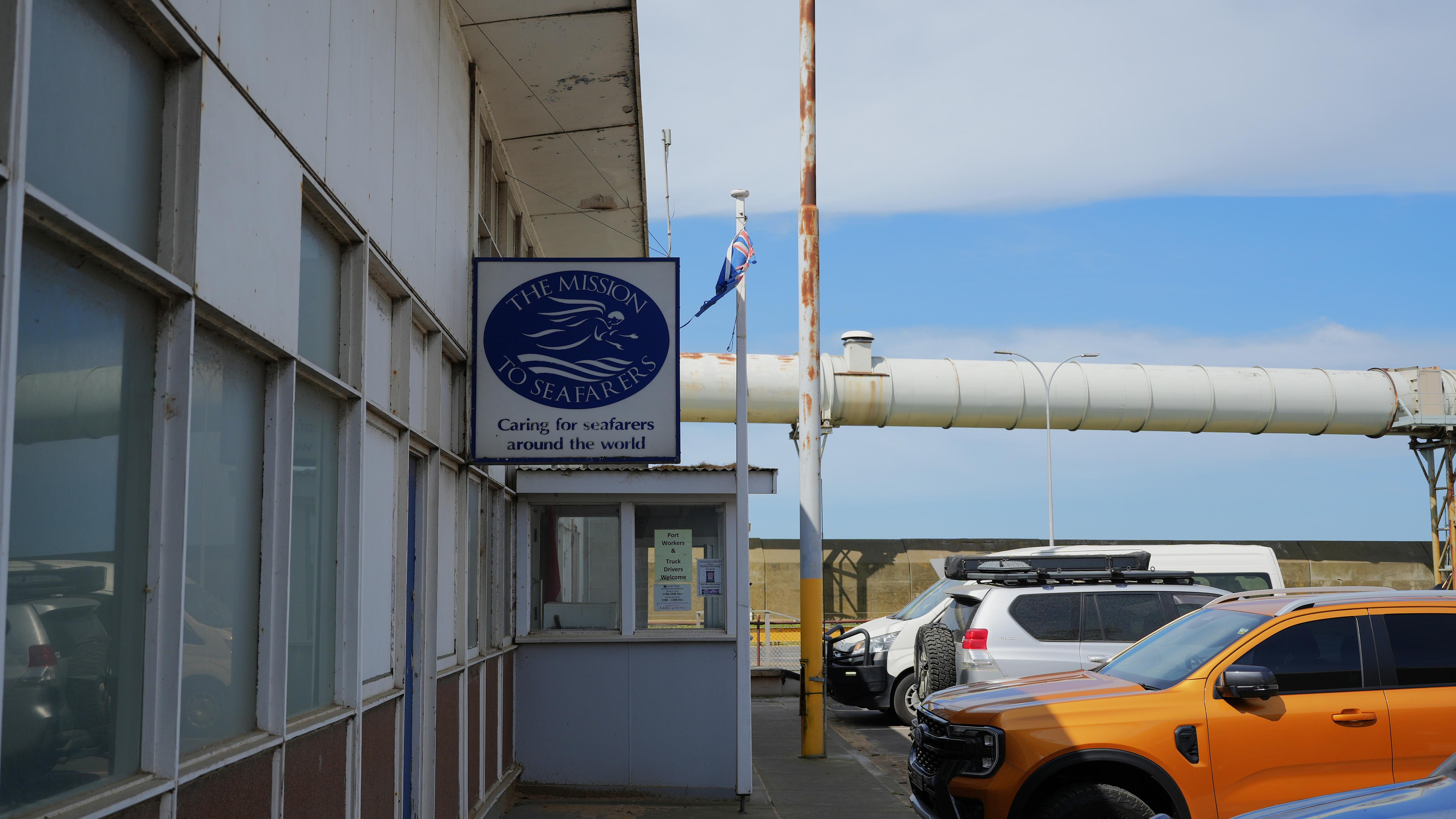 The outside of a one-storey building, a worn down sign has the Mission to Seafarers logo and an Australian flag flies above.