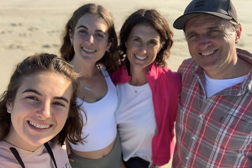 Three women with brown hair and a man with a cap on the beach smiling