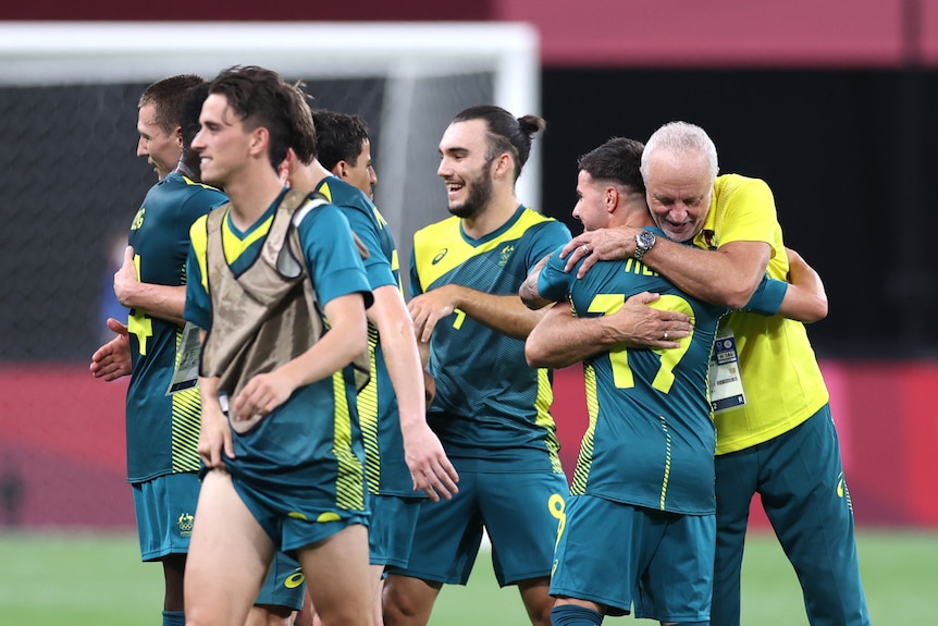 Olyroos coach Graham Arnold hugs one of his players as his teammates smile after a big win at the Olympics.