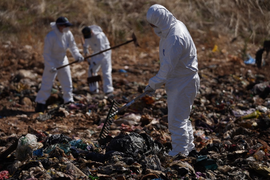 An officer wearing protective gear rakes through rubbish.