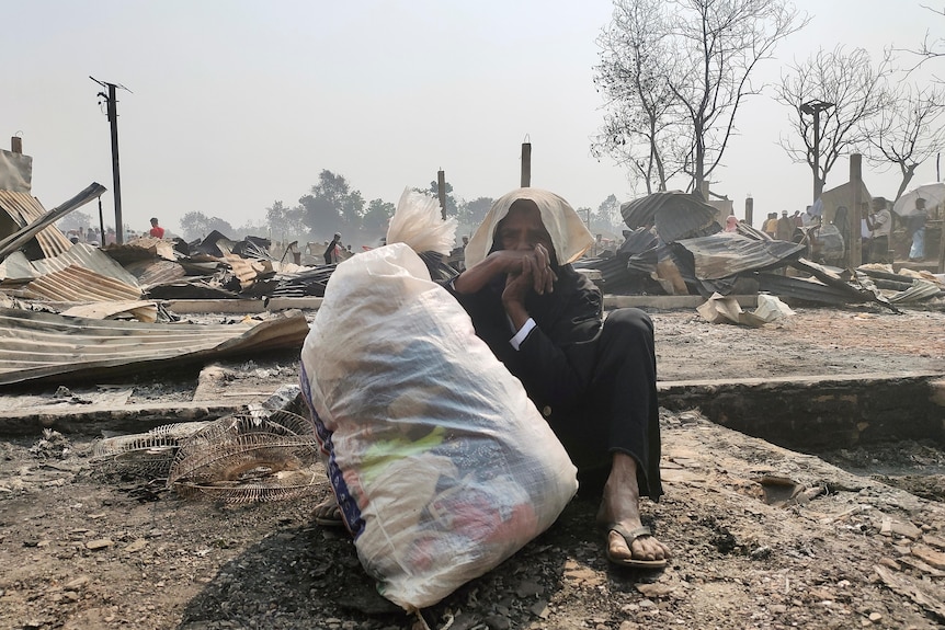Une vieille femme asiatique est assise avec un sac blanc d'objets en face du village brûlé.