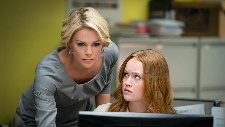 Seated woman glances up to a woman standing close with short blonde hair and serious expression focused on computer screen.