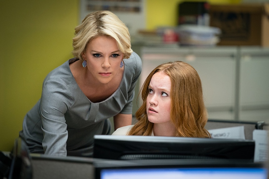 Seated woman glances up to a woman standing close with short blonde hair and serious expression focused on computer screen.