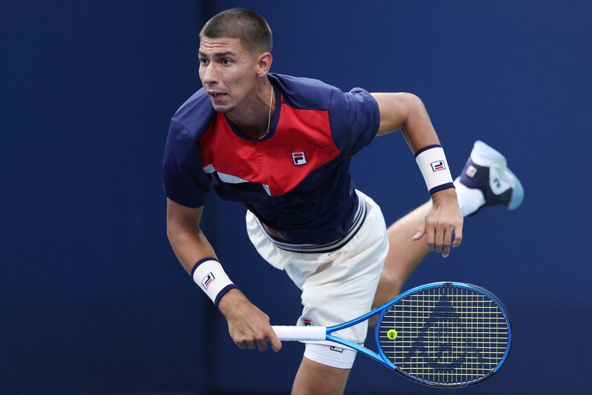 A male tennis player, weairng a red white, dark blue shirt, and white shorts, follows through a serve.