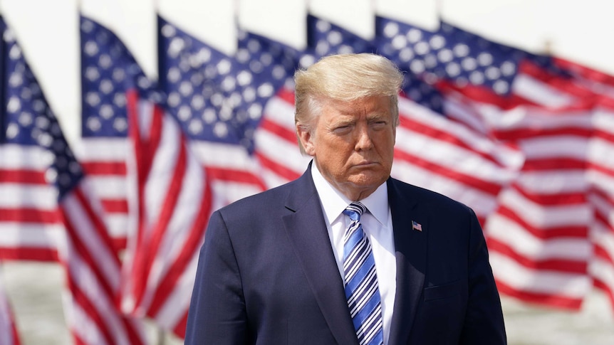 Donald Trump looking serious in front of a row of American flags