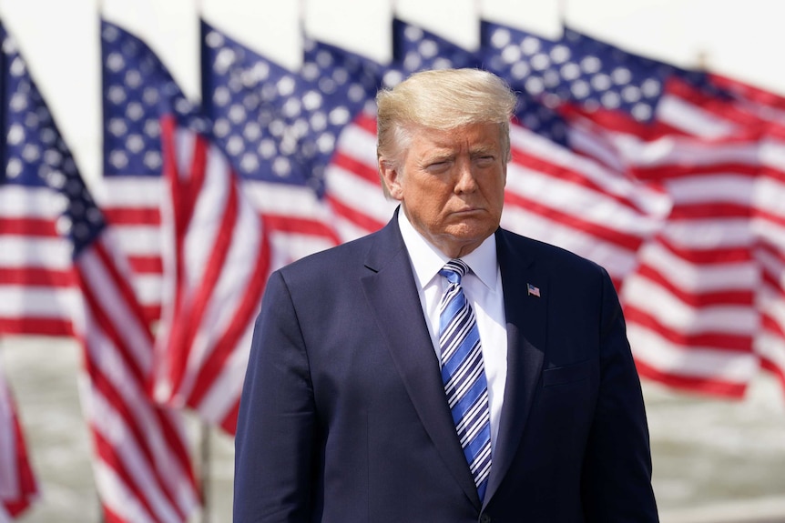 Donald Trump looking serious in front of a row of American flags