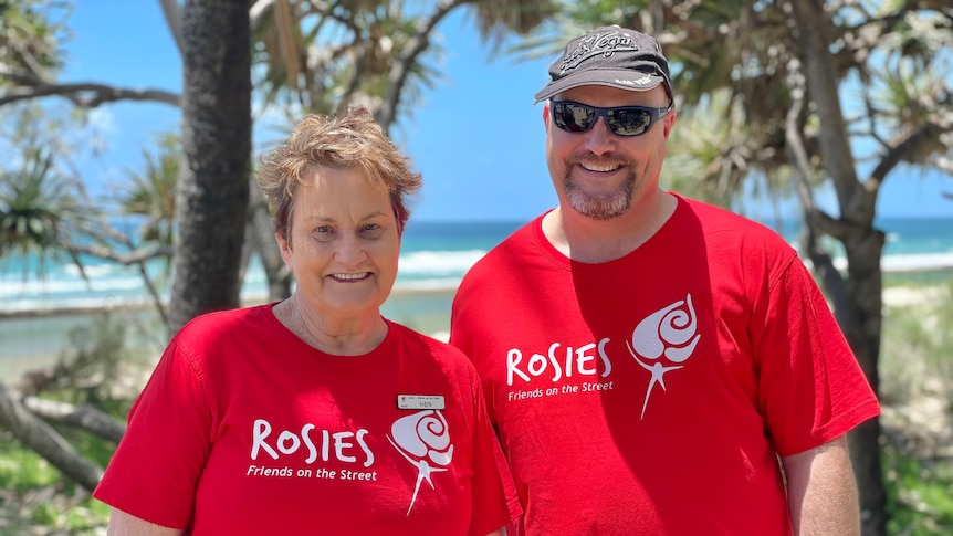 A woman with short hair and a taller man wearing hat and sunglasses at the beach, both wearing red t-shirts that read "Rosies"