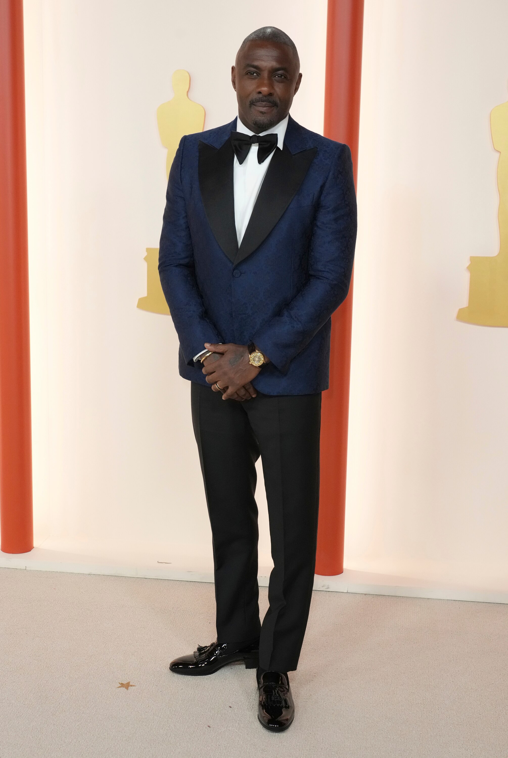 Idris Elba wearing a dark blue suit jacket with a white button-up shirt, black trousers and a black bow tie. 