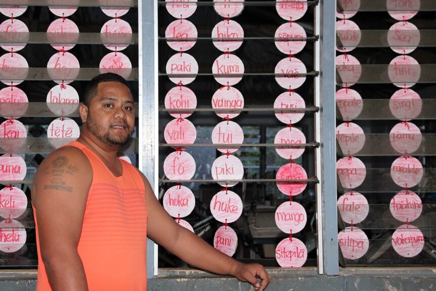 Tevita, wearing a singlet, stands outside his classroom, the children's art visible inside the window