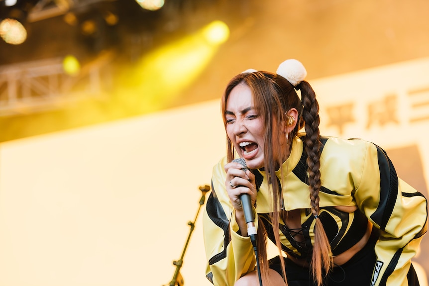 A Taiwanese Australian woman with braided copper hair wears a black and yellow jacket and sings on stage under yellow lights.