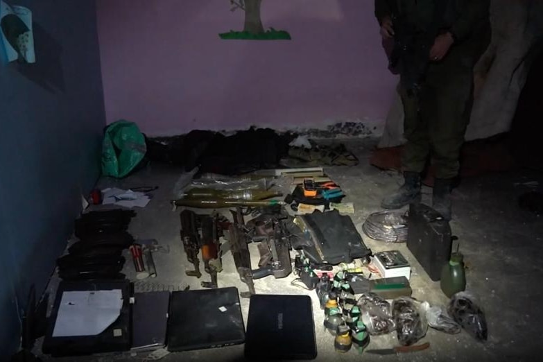 Rifles, grenades, laptops and other devices are laid out on a bare floor in a dark room.