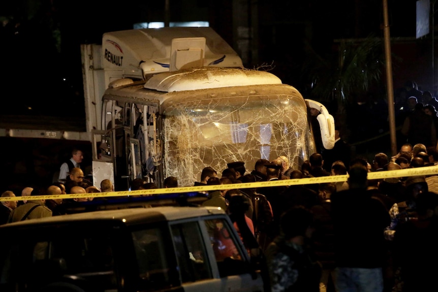 A crowd of officials gather near the front of a mini bus with a shattered windscreen and blown out sides.