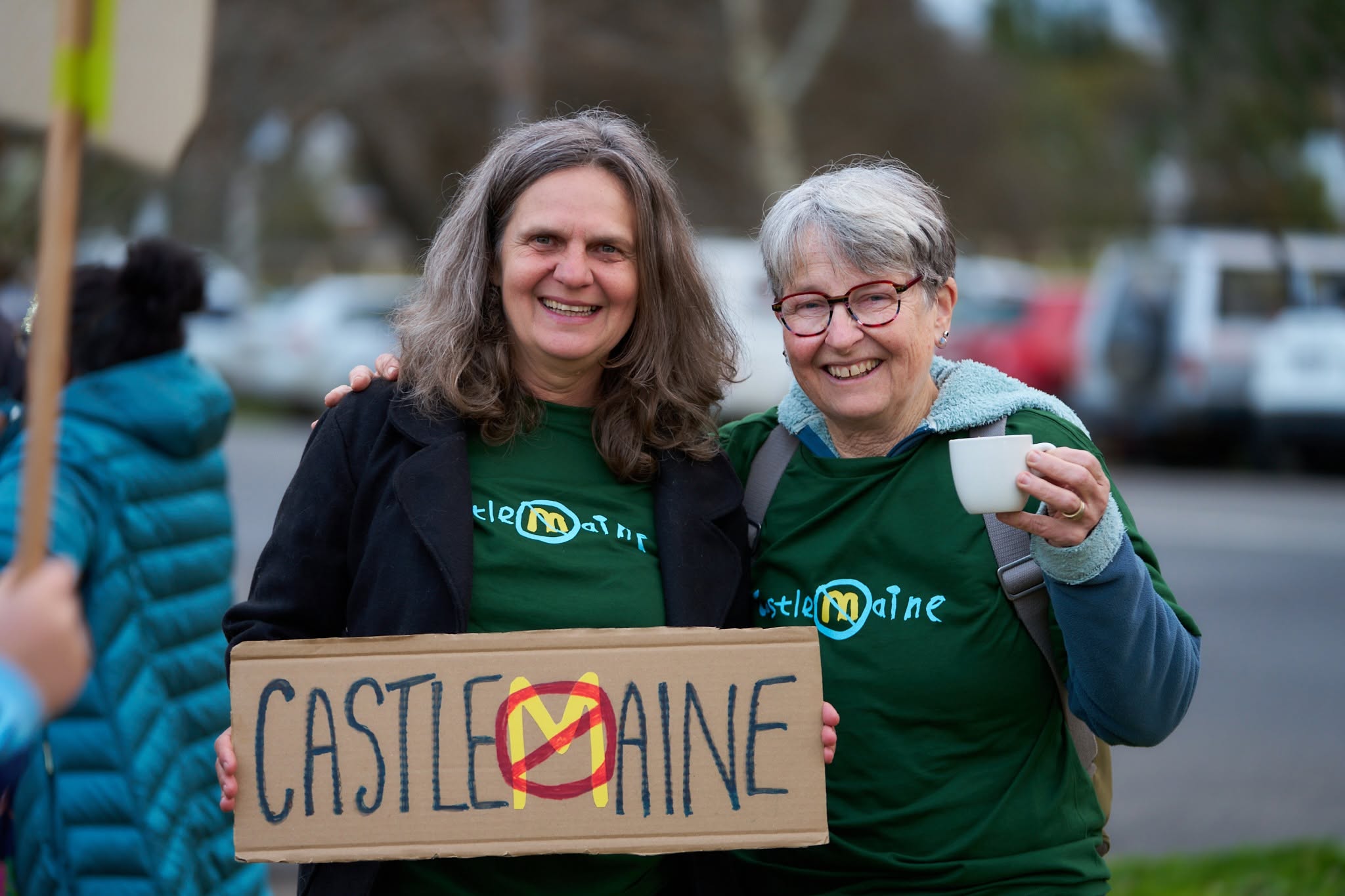 Photo of two women at a protest, holding up a sign that says Castlemaine and the M has a slash through it