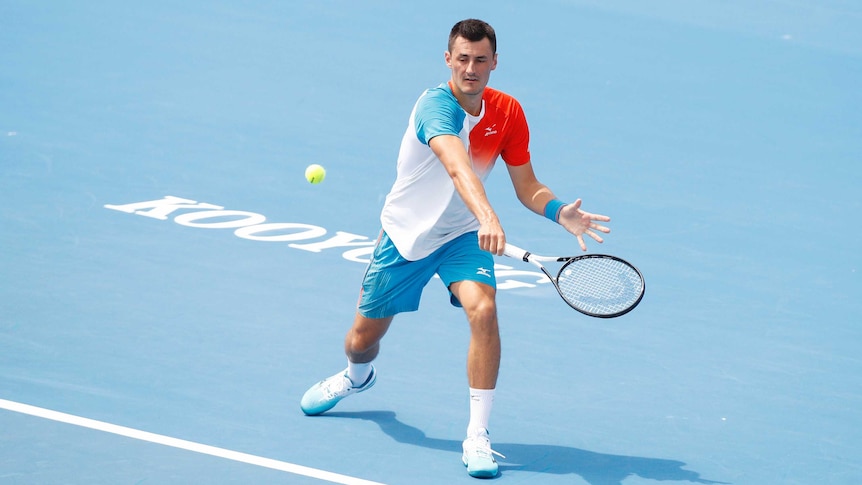 A tennis player hits a back-hand shot with his racquet in his right hand