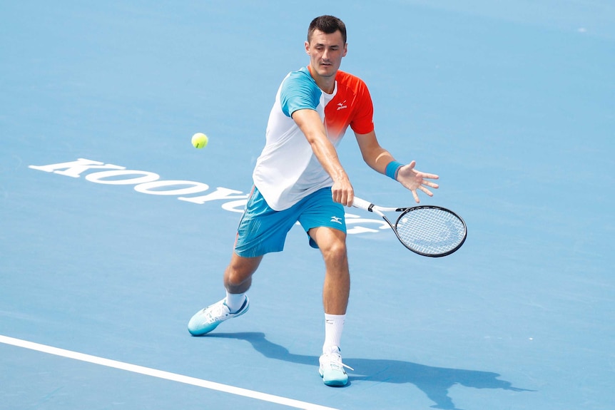 A tennis player hits a back-hand shot with his racquet in his right hand